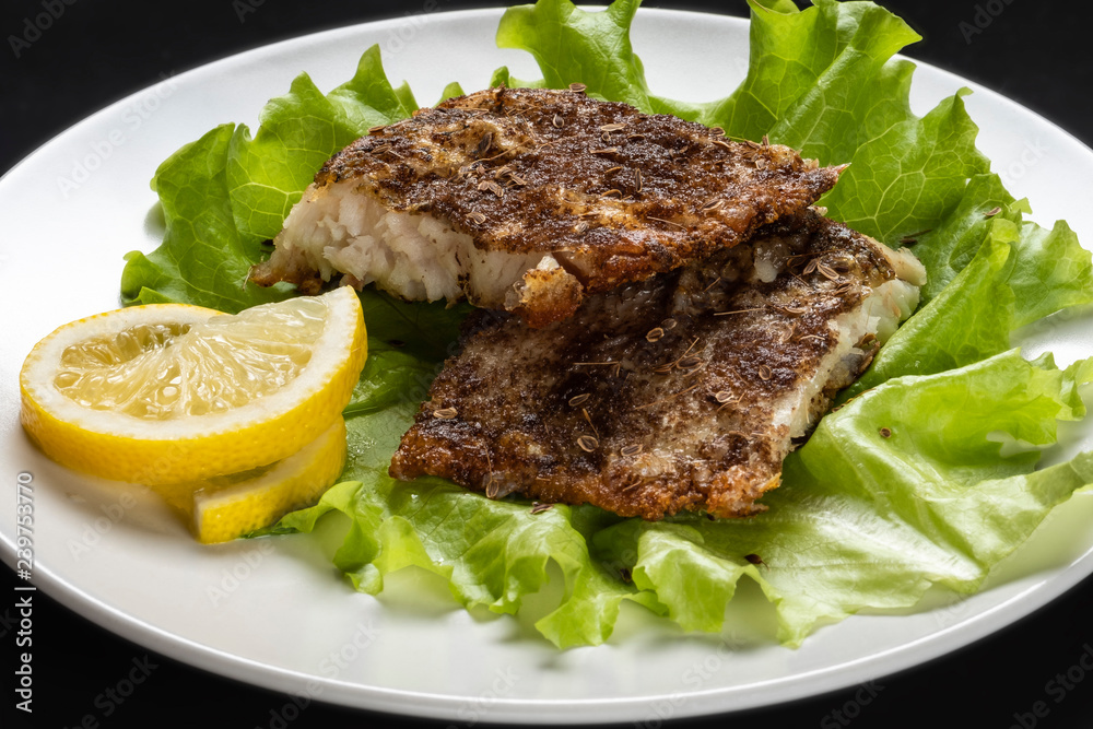 Two pieces of grilled fish fillet with the fresh leafs of lettuce and lemon on white plate on black background. Close up. Two pieces of grilled fish fillet with the fresh leafs of lettuce and lemon on white plate on black background. Close up.