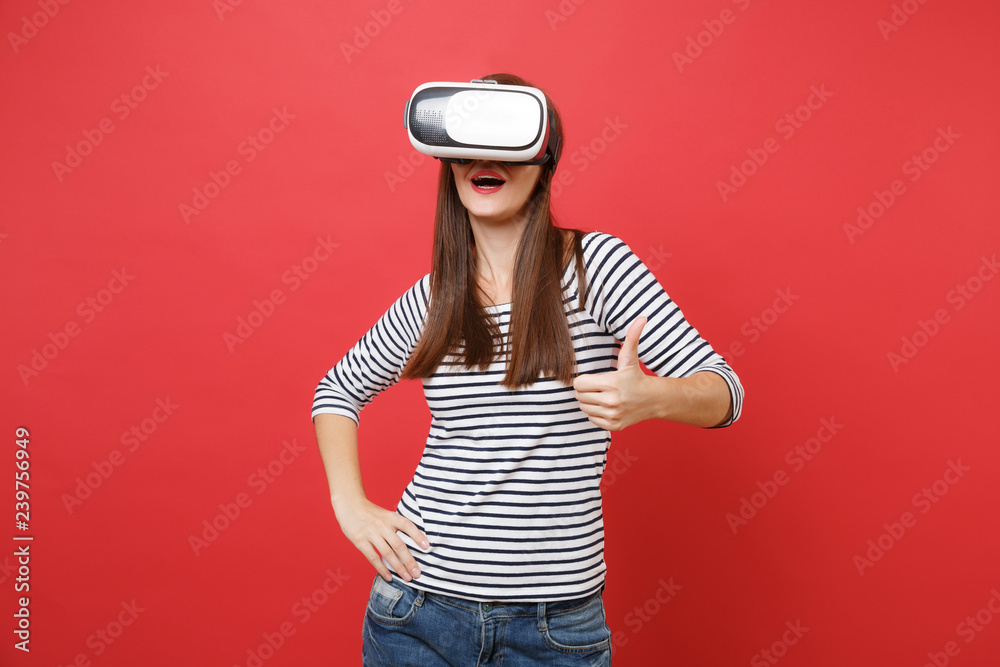 Portrait of young woman in striped clothes, virtual reality glasses standing and showing thumb up isolated on bright red wall background. People sincere emotions lifestyle concept. Mock up copy space.