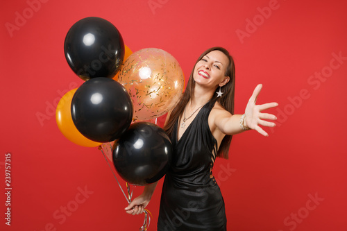 Smiling beautiful young woman in black dress pointing hand on camera holding air balloons, celebrating isolated on red background. Valentine's Day Happy New Year birthday mockup holiday party concept. © ViDi Studio
