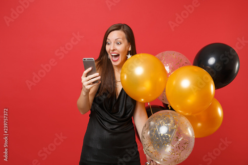 Excited happy young girl in little black dress holding air balloons using mobile phone while celebrating isolated on red background. Women's Day, Happy New Year, birthday mockup holiday party concept. © ViDi Studio