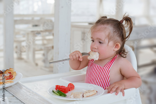A little smiling toddler girl is sitting at a table in a summer cafe in the seaside resort.