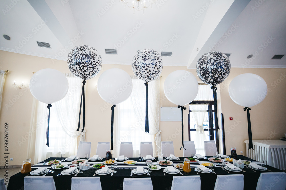 Festive dinner table decor. White and black balloons hang over the restaurant table served with delicious food - obrazy, fototapety, plakaty 