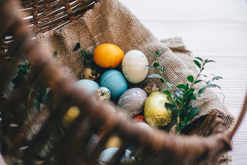Stylish easter eggs on cloth in rustic basket on white wooden background. Easter hunt concept. Modern easter eggs painted with natural dye and green buxus branches. Happy Easter