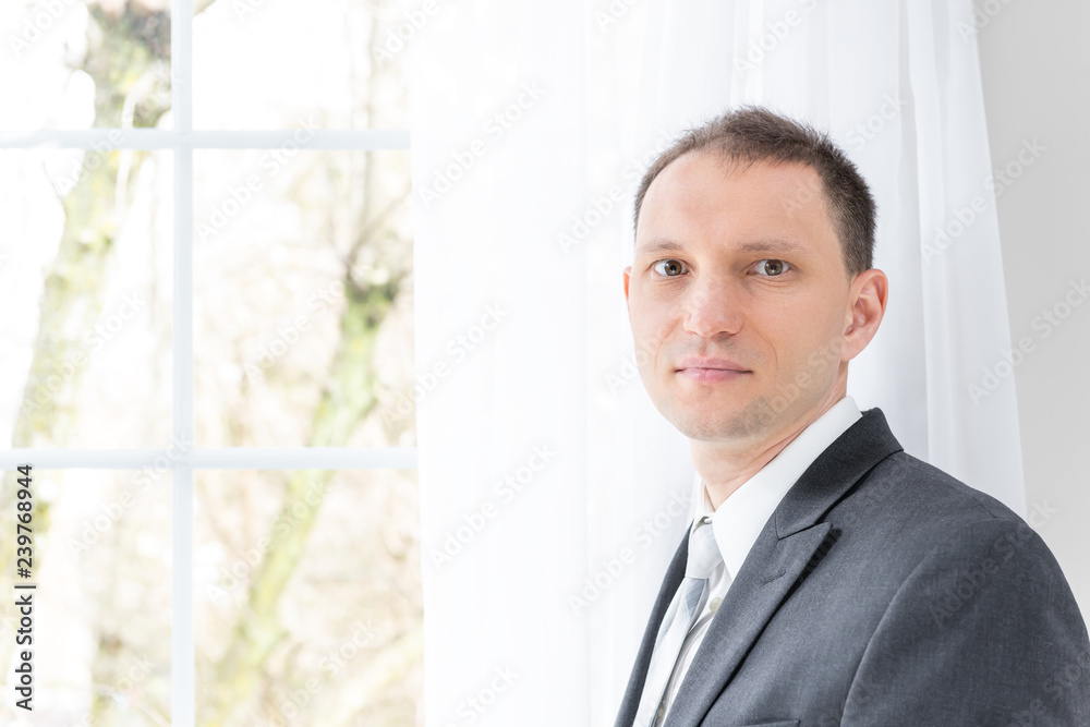 Closeup of young man, male businessman in business gray, grey suit, tie, bridegroom, standing by window in home, house room, white curtains, looking at camera, thinking Closeup of young man, male businessman in business gray, grey suit, tie, bridegroom, standing by window in home, house room, white curtains, looking at camera, thinking