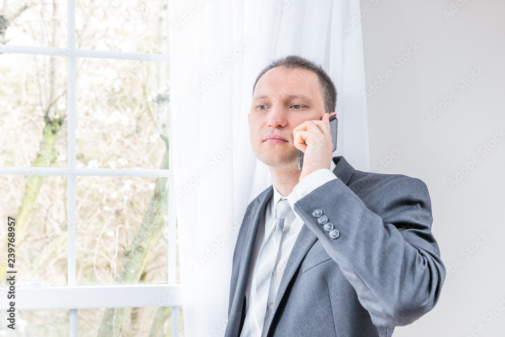Closeup of young man, male businessman in business gray, grey suit, tie, bridegroom, standing by window in home, house room, white curtains, talking on phone, thinking Closeup of young man, male businessman in business gray, grey suit, tie, bridegroom, standing by window in home, house room, white curtains, talking on phone, thinking