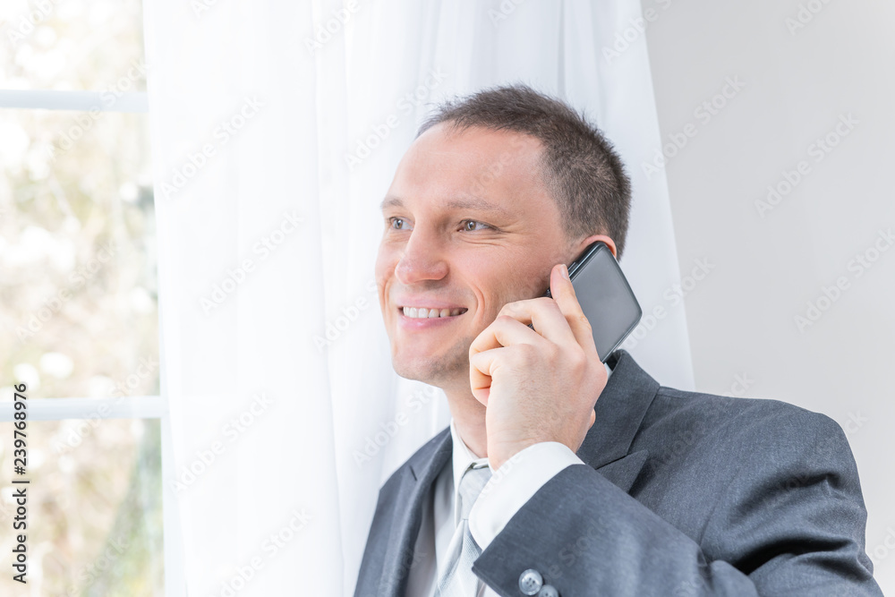 Closeup of young, happy, smiling, man, male businessman in business gray suit, tie, bridegroom, standing by window in home, house room, white curtains, talking on phone Closeup of young, happy, smiling, man, male businessman in business gray suit, tie, bridegroom, standing by window in home, house room, white curtains, talking on phone