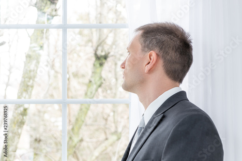 Closeup profile, side of young man, male businessman in business gray, grey suit, tie, bridegroom, standing in home, house, apartment room, white curtains, looking through window on view, thinking