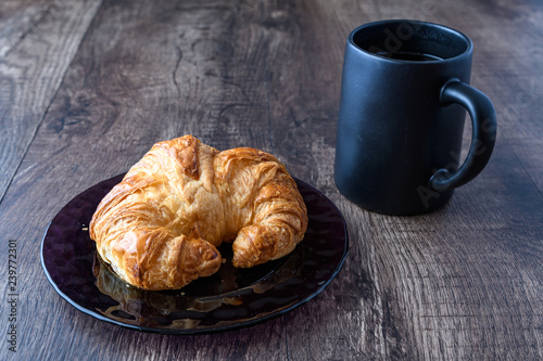 Fresh croissant on a purple glass plate on a wood table top with black coffee cup, side view