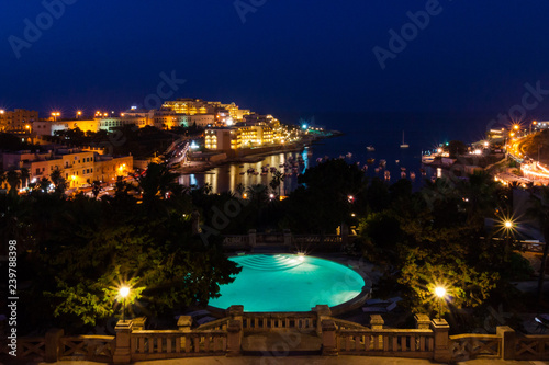 View of St. George's Bay seafront lights by night, with a blue luxury swimming pool and boats and yachts anchored. St Julian's (San Giljan), Central Region, Malta. Evening in Paceville district