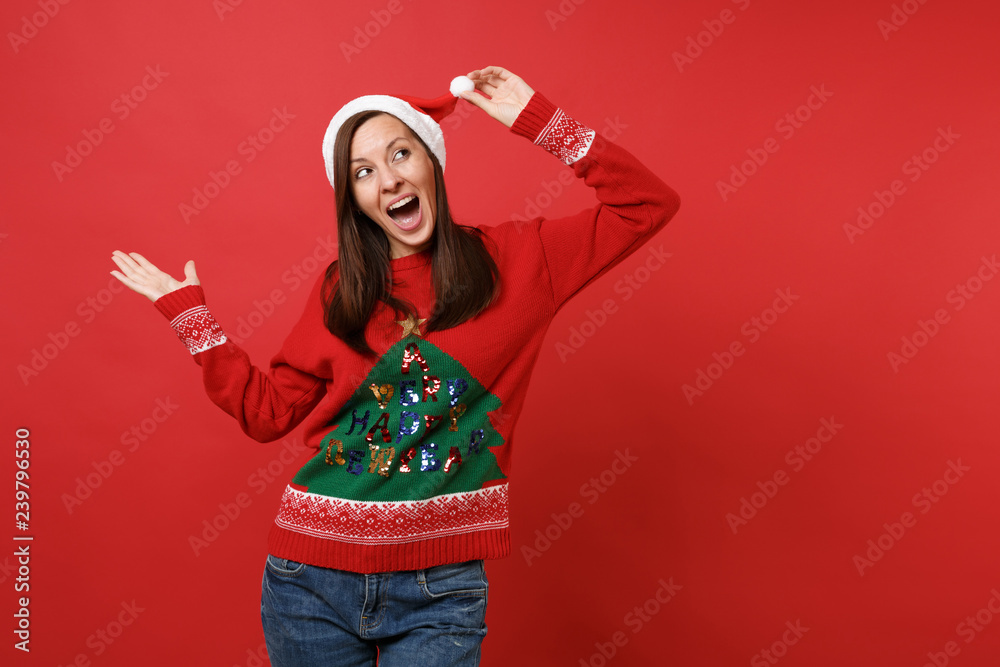 Surprised young Santa girl keeping hand on Christmas hat and mouth wide open, pointing hand aside isolated on red background. Happy New Year 2019 celebration holiday party concept. Mock up copy space. Surprised young Santa girl keeping hand on Christmas hat and mouth wide open, pointing hand aside isolated on red background. Happy New Year 2019 celebration holiday party concept. Mock up copy space.