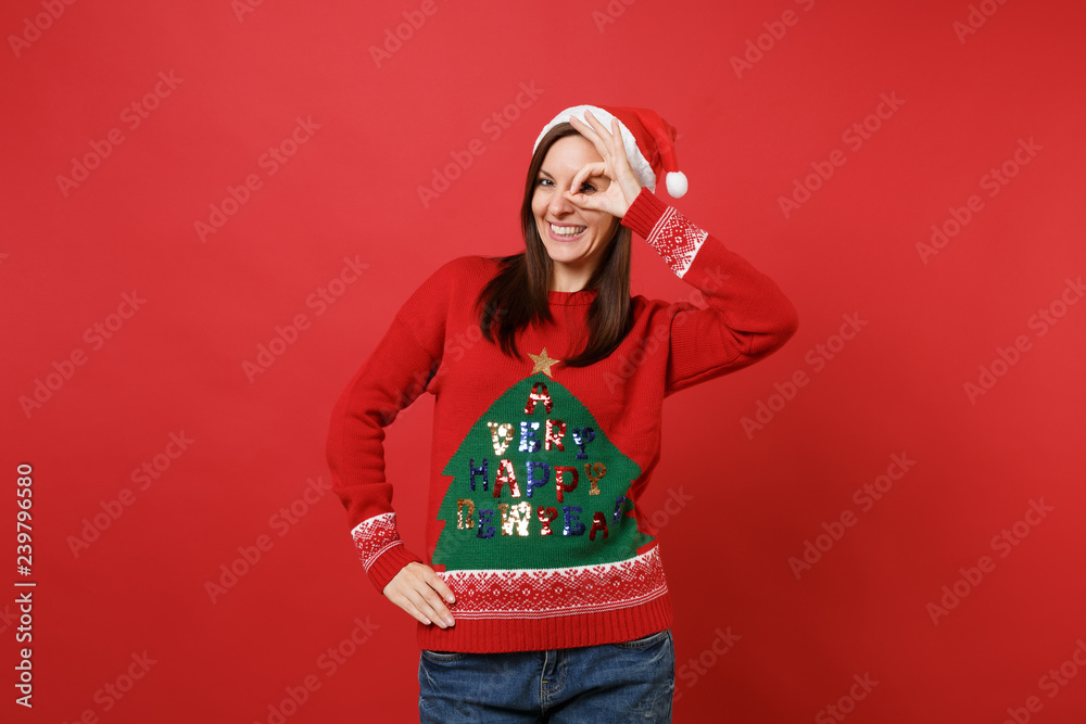 Smiling young Santa girl in knitted sweater and Christmas hat showing OK gesture near eyes isolated on bright red background. Happy New Year 2019 celebration holiday party concept. Mock up copy space. Smiling young Santa girl in knitted sweater and Christmas hat showing OK gesture near eyes isolated on bright red background. Happy New Year 2019 celebration holiday party concept. Mock up copy space.