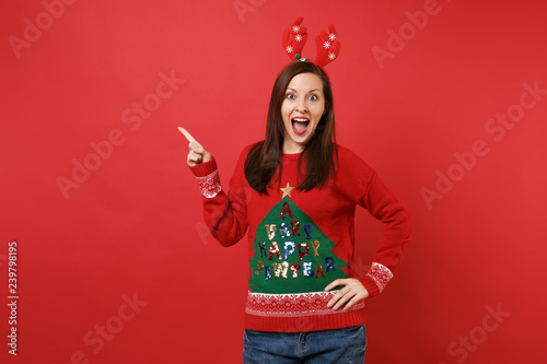 Amazed Santa girl in fun decorative deer horns pointing index finger aside, keeping mouth wide open looking surprised isolated on red background. Happy New Year 2019 celebration holiday party concept.