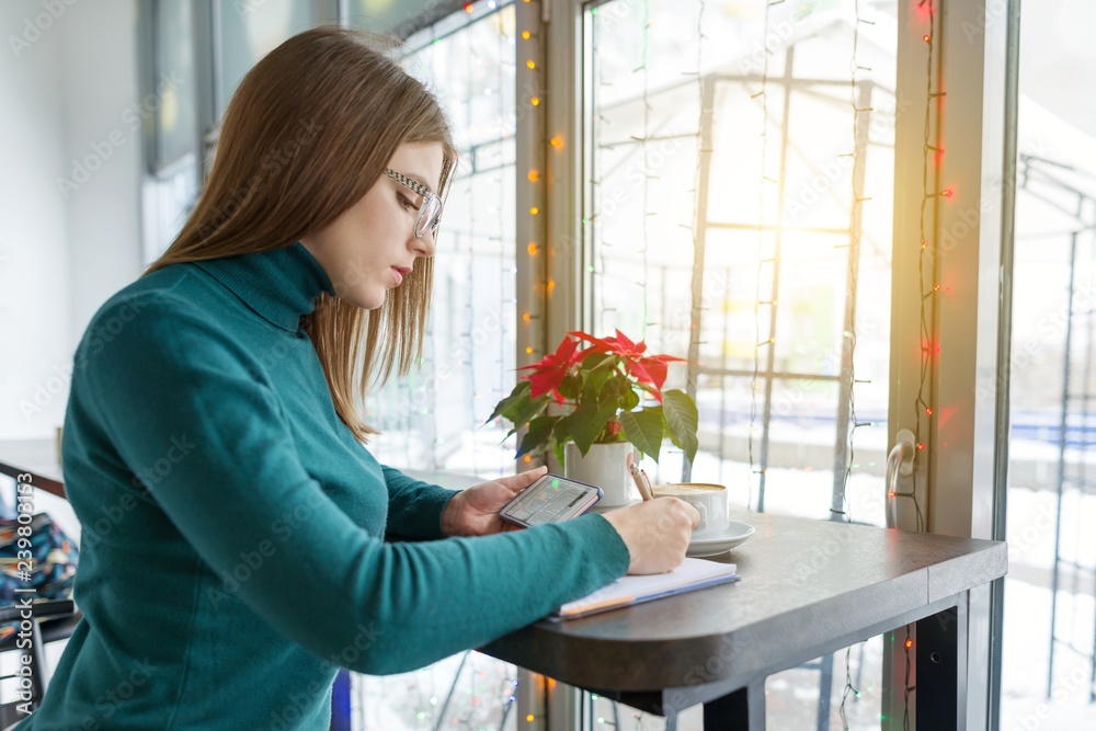 Winter portrait of student girl studying in cafe sitting with cup of coffee. Young woman writing in notebook using smartphone for learning Winter portrait of student girl studying in cafe sitting with cup of coffee. Young woman writing in notebook using smartphone for learning
