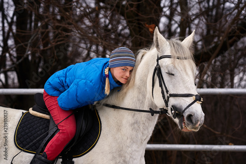 Young woman rider in a blue blazer and sporting a cap for a walk on a white horse on a cloudy winter day.