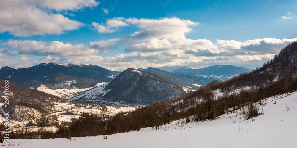 panorama of winter countryside in mountains. wonderful sunny day with gorgeous cloudscape. dark forest on the snow covered hills. village down in the distant valley. faraway ridge with snowy tops