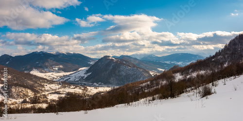 panorama of winter countryside in mountains. wonderful sunny day with gorgeous cloudscape. dark forest on the snow covered hills. village down in the distant valley. faraway ridge with snowy tops © Pellinni