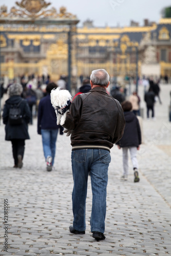 a white dog in the arms of a man in a brown leather jacket walks outside