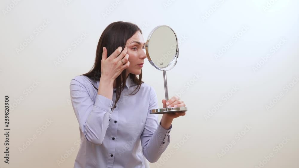 A pretty brunette is looking into the makeup mirror, adjusts her eyebrows, touches the skin under eyes and right temple A pretty brunette is looking into the makeup mirror, adjusts her eyebrows, touches the skin under eyes and right temple