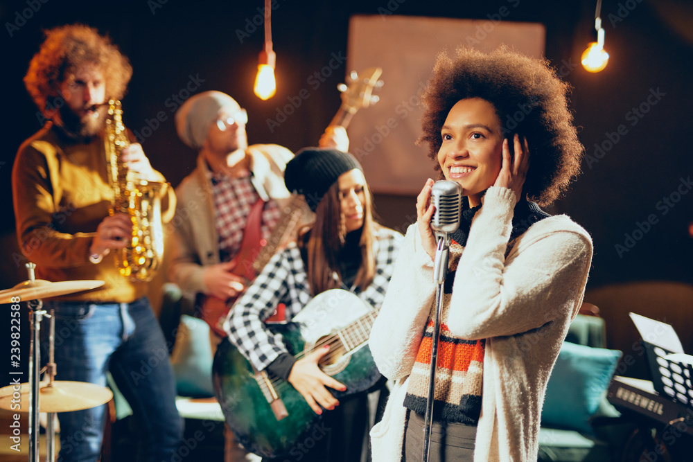 Naklejka premium Mixed race woman singing. In background band playing instruments. Home studio interior.