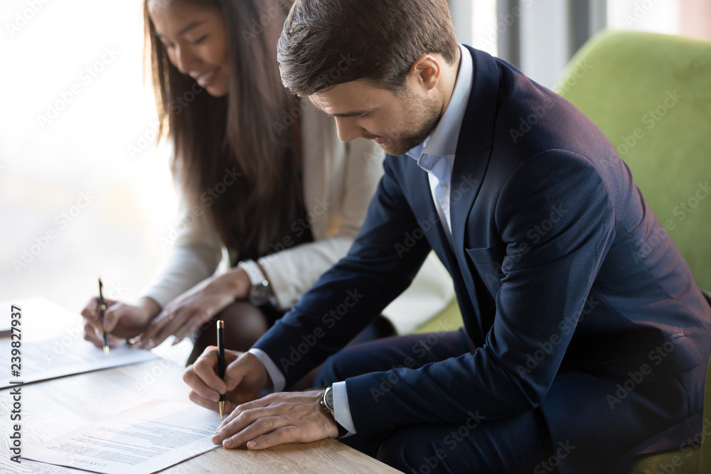 Fototapeta premium Positive asian businesswoman sitting on couch with business partner, focus on caucasian businessman, people signing contract agreement document. Successful negotiations, trust and partnership concept