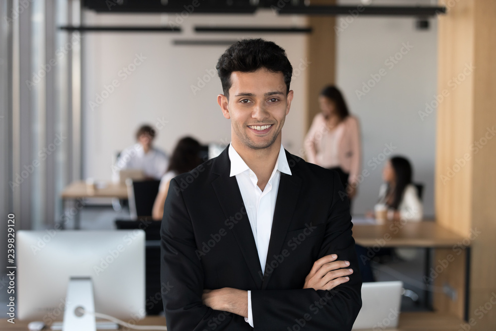 Head shot of confident millennial middle eastern ethnicity office employee or team leader wearing formal suit standing with arms crossed on chest posing looking at camera with coworkers on background. Head shot of confident millennial middle eastern ethnicity office employee or team leader wearing formal suit standing with arms crossed on chest posing looking at camera with coworkers on background.