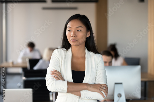 Head shot of serious asian woman stands in coworking office with hands crossed feels dissatisfaction. Pensive businesswoman company employee having difficulties at work thinking about problem solving
