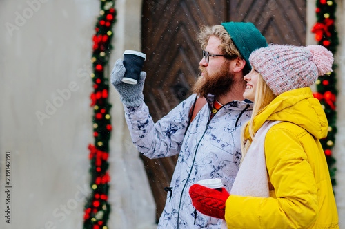 Winter holidays, travel, relarions concept: happy smiling hipster couple in bright warm outwear walking, looking aside, holding cups with hot drink, posing in street. Snowfall. Copy space photo