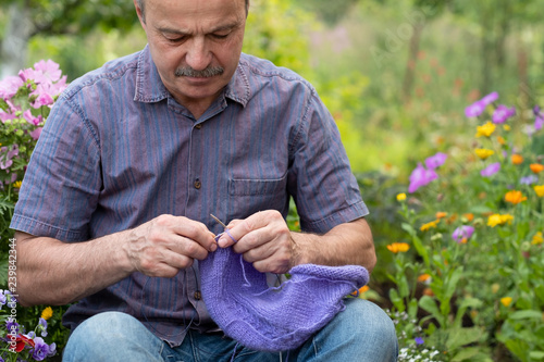 Old handsome man sitting at his summer garden with the knitting needles. A man learn to knit needles sweater. Hobby at senior age photo