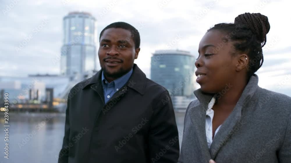Medium shot of African man and woman walking along embankment in urban area and having conversation Medium shot of African man and woman walking along embankment in urban area and having conversation