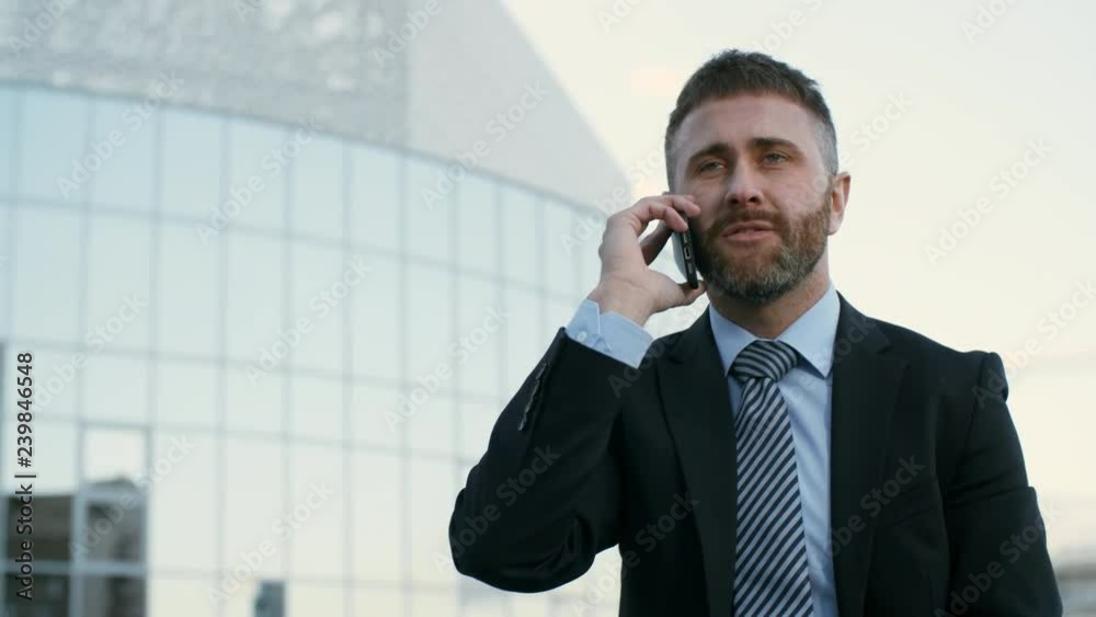 Pan shot of Caucasian businessman sitting outdoors on bench, talking on phone and laughing Pan shot of Caucasian businessman sitting outdoors on bench, talking on phone and laughing