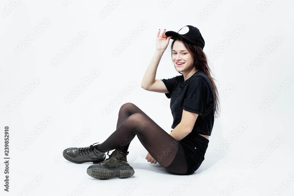 Smiling young dark-haired girl dressed in a black top, shorts, tights and  cap poses sitting on the floor on the white background in the studio Photos  | Adobe Stock