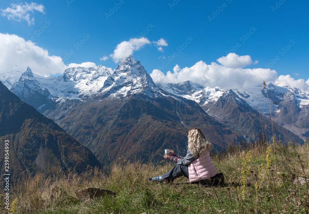 Fototapeta premium woman traveler drinks coffee with a view of the mountain landscape. A young tourist woman drinks a hot drink from a cup and enjoys the scenery in the mountains. Trekking concept.
