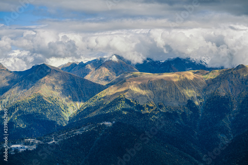 Mountain landscape. Warm autumn Sunny day. The slopes of the mountains covered with autumn woods. Clouds above mountain peaks. The snow on the tops