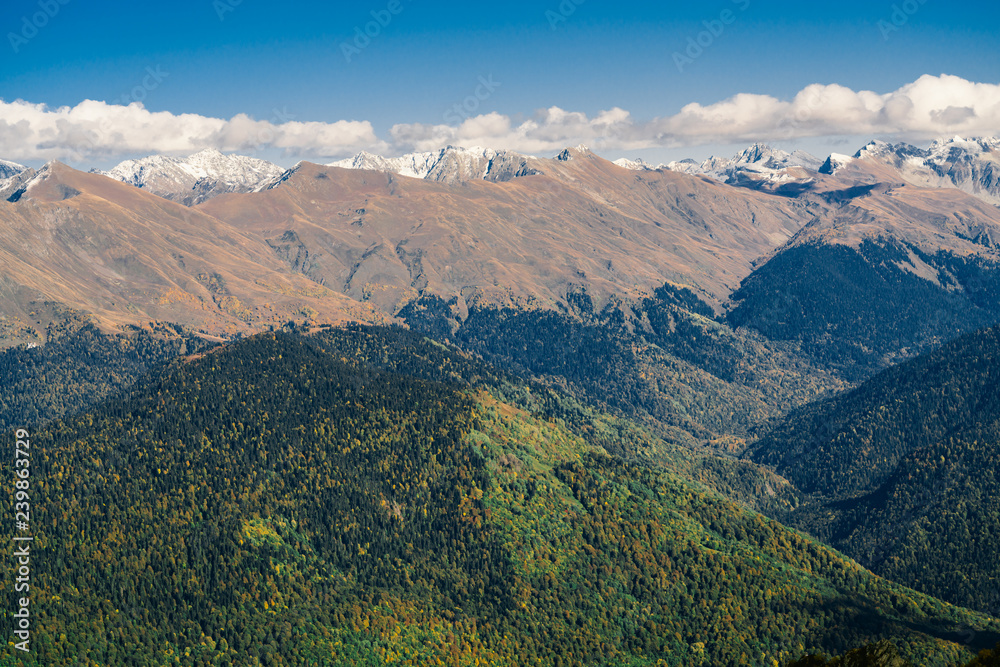 Mountain landscape. Warm autumn Sunny day. The slopes of the mountains covered with autumn woods. Clouds above mountain peaks. The snow on the tops