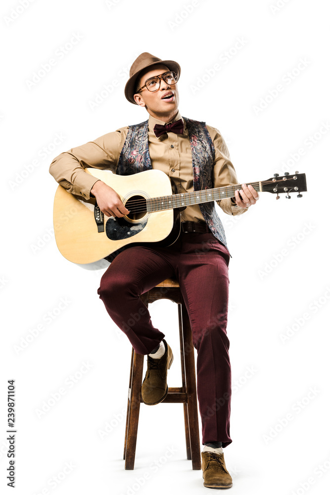 smiling mixed race male musician in hat and eyeglasses playing on acoustic guitar while sitting on chair isolated on white smiling mixed race male musician in hat and eyeglasses playing on acoustic guitar while sitting on chair isolated on white