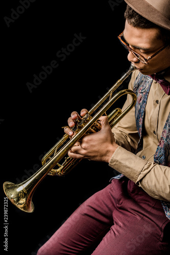mixed race male jazzman in hat and eyeglasses playing on trumpet isolated on black