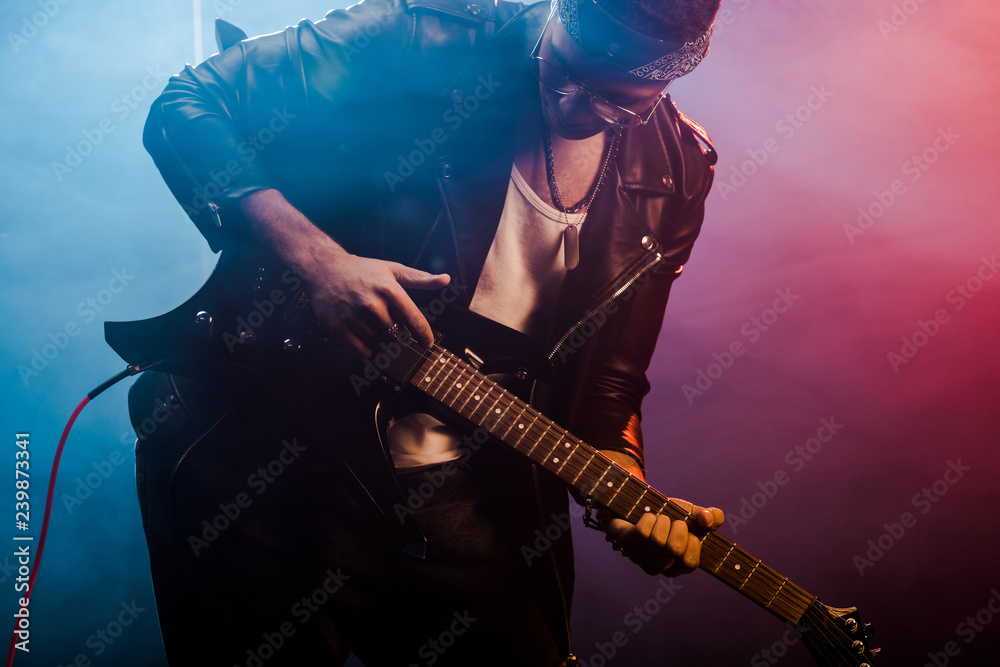 young male rocker in leather jacket performing on electric guitar on stage with smoke and dramatic lighting