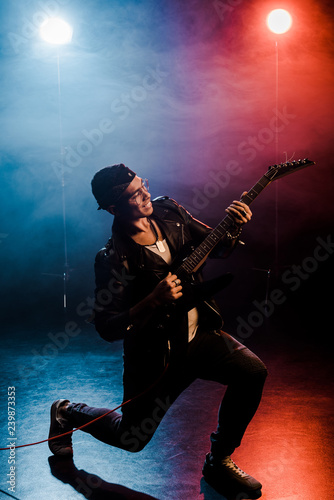 smiling male rock star in leather jacket performing on electric guitar on stage with smoke and dramatic lighting