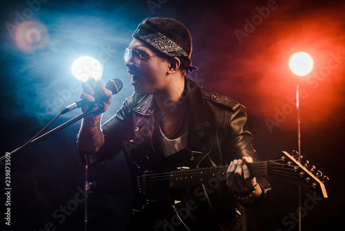 emotional male musician singing in microphone and playing on electric guitar on stage during rock concert