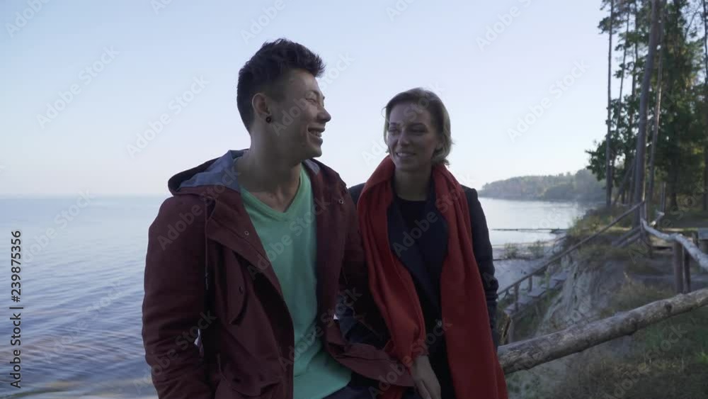 Young smiling interracial couple in casual wear sitting by the cliff on a background of pine trees and a lake. Multi ethnic couple in love enjoying a date, smiling happily and talking on the Young smiling interracial couple in casual wear sitting by the cliff on a background of pine trees and a lake. Multi ethnic couple in love enjoying a date, smiling happily and talking on the