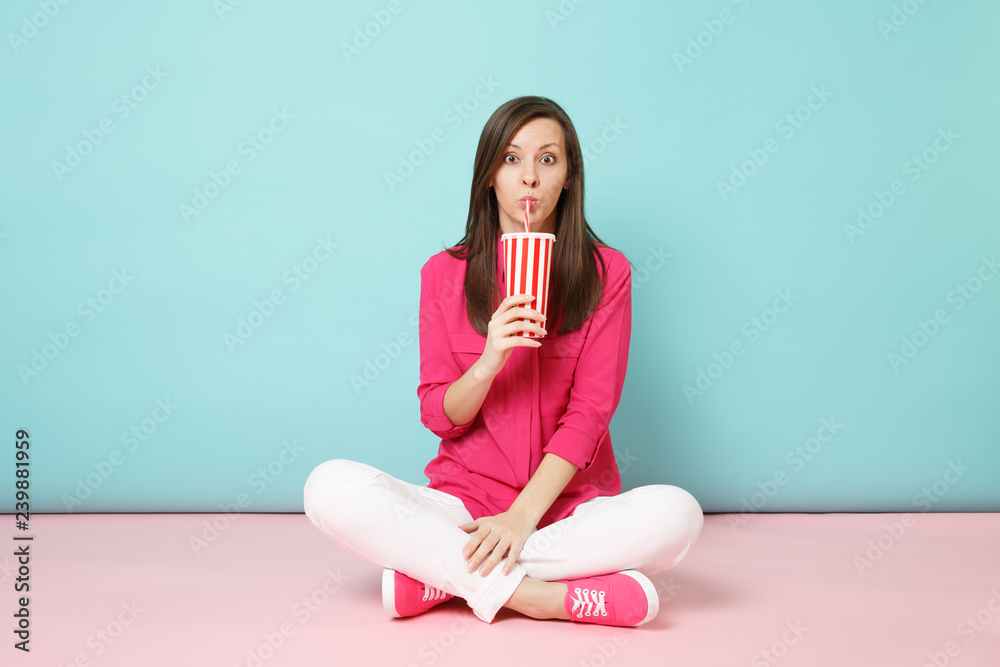 Full length portrait woman in rose shirt blouse white pants sitting on floor hold cup of soda isolated on bright pink blue pastel wall background studio. Fashion lifestyle concept. Mock up copy space. Full length portrait woman in rose shirt blouse white pants sitting on floor hold cup of soda isolated on bright pink blue pastel wall background studio. Fashion lifestyle concept. Mock up copy space.