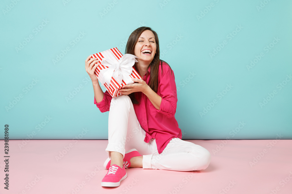 Full length portrait woman in rose shirt blouse white pants sitting on floor hold present box isolated on bright pink blue pastel wall background studio. Fashion lifestyle concept. Mock up copy space.
