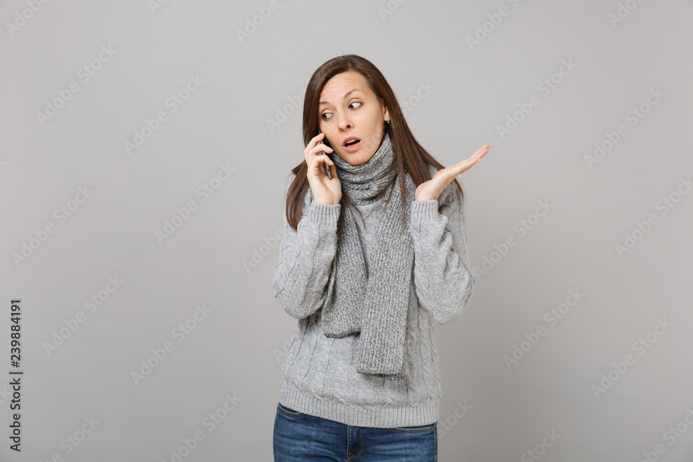 Confused young woman in gray sweater scarf spreading hands talking on mobile phone isolated on grey wall background. Healthy fashion lifestyle people emotions, cold season concept. Mock up copy space. Confused young woman in gray sweater scarf spreading hands talking on mobile phone isolated on grey wall background. Healthy fashion lifestyle people emotions, cold season concept. Mock up copy space.