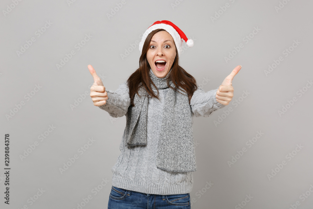 Excited Santa girl in gray sweater scarf Christmas hat keeping mouth wide open showing thumbs up isolated on grey background. Happy New Year 2019 celebration holiday party concept. Mock up copy space. Excited Santa girl in gray sweater scarf Christmas hat keeping mouth wide open showing thumbs up isolated on grey background. Happy New Year 2019 celebration holiday party concept. Mock up copy space.