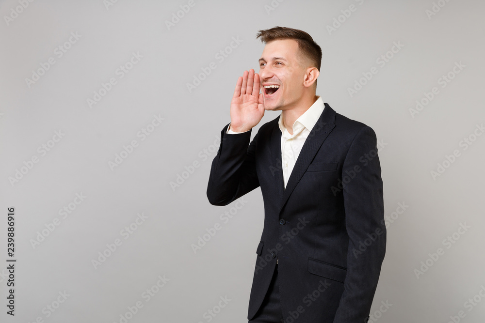 Portrait of joyful young business man in classic black suit and shirt screaming with hand gesture isolated on grey background in studio. Achievement career wealth business concept. Mock up copy space. Portrait of joyful young business man in classic black suit and shirt screaming with hand gesture isolated on grey background in studio. Achievement career wealth business concept. Mock up copy space.