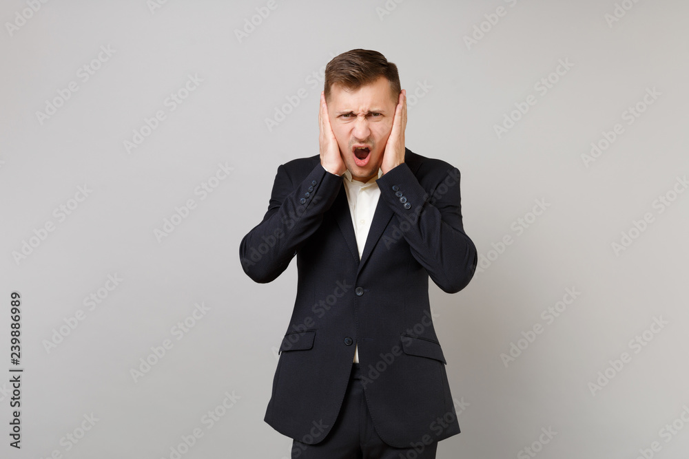 Preoccupied young business man in classic black suit, shirt covering ears with hands, screaming isolated on grey background in studio. Achievement career wealth business concept. Mock up copy space. Preoccupied young business man in classic black suit, shirt covering ears with hands, screaming isolated on grey background in studio. Achievement career wealth business concept. Mock up copy space.