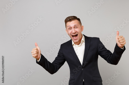 Portrait of happy young business man in classic black suit, shirt showing thumb up screaming isolated on grey wall background in studio. Achievement career wealth business concept. Mock up copy space.