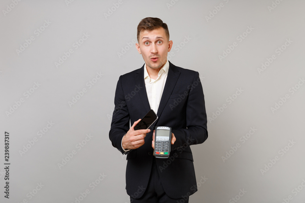 Amazed business man holding wireless modern bank payment terminal to process, acquire credit card payments, mobile phone isolated on grey wall background. Achievement career wealth business concept. Amazed business man holding wireless modern bank payment terminal to process, acquire credit card payments, mobile phone isolated on grey wall background. Achievement career wealth business concept.
