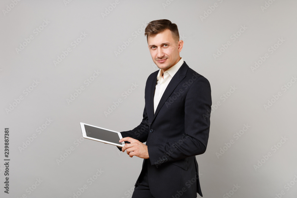 Handsome young business man in classic black suit holding, using tablet pc computer with blank empty screen isolated on grey background. Achievement career wealth business concept. Mock up copy space. Handsome young business man in classic black suit holding, using tablet pc computer with blank empty screen isolated on grey background. Achievement career wealth business concept. Mock up copy space.
