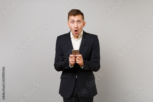 Shocked young business man keeping mouth open wide, looking surprised using mobile phone typing sms message isolated on grey background. Achievement career wealth business concept. Mock up copy space.